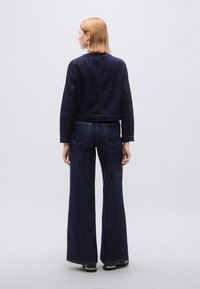 Σκούρο μπλε τζιν cropped μπουφάν με μακριά μανίκια, συνδυασμένο με σκούρο μπλε τζιν παντελόνι φαρδιάς γραμμής. Και τα δύο κομμάτια έχουν ελάχιστες ραφές και λεπτομέρειες στις τσέπες.