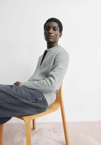 Hellgraues Sweatshirt mit Reißverschluss vorne und gerippten Bündchen, getragen über einem schwarzen Oberteil; kombiniert mit dunklen Denim-Hosen; sitzt auf einem Stuhl aus hellem Holz.