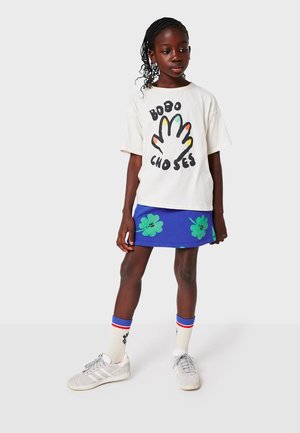 T-shirt blanc avec un graphisme de main coloré, associé à une jupe bleue ornée de motifs floraux verts. Porté avec des baskets grises et des chaussettes rayées.