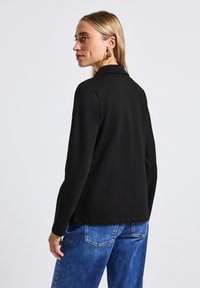 Černá bunda s dlouhým rukávem, šitá s límcem, hladkým povrchem a bočními rozparky, kombinovaná s modrými džíny. Minimalistický design a strukturovaný střih.