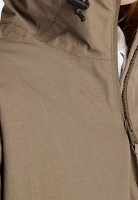 Veste imperméable beige avec un col montant, une capuche à cordon de serrage réglable et une fermeture éclair latérale. Elle présente une texture lisse et des coutures discrètes.