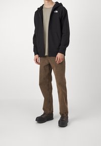 Felpa nera con zip e tasche a marsupio, indossata sopra una t-shirt grigia girocollo, abbinata a pantaloni marroni e scarpe nere con lacci.