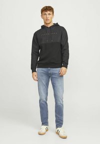 Sudadera negra y gris con un logo frontal, cordones ajustables y puños acanalados, combinada con jeans azul claro y zapatillas blancas.
