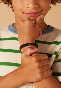 Zwarte gevlochten polsband om de pols van een kind; de achtergrond toont een gestreepte trui in wit met groene en blauwe accenten. Huidtint is bruin.
