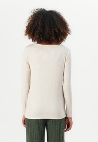 Kvinna med lockigt hår som bär en beige långärmad ribbad topp och gröna ribbade byxor, stående vänd mot en enkel vit bakgrund.