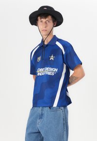 Blue short-sleeve polo with wave pattern, white accents, and "Obey Design Industries" logo. Worn with light blue denim shorts and a black bucket hat.