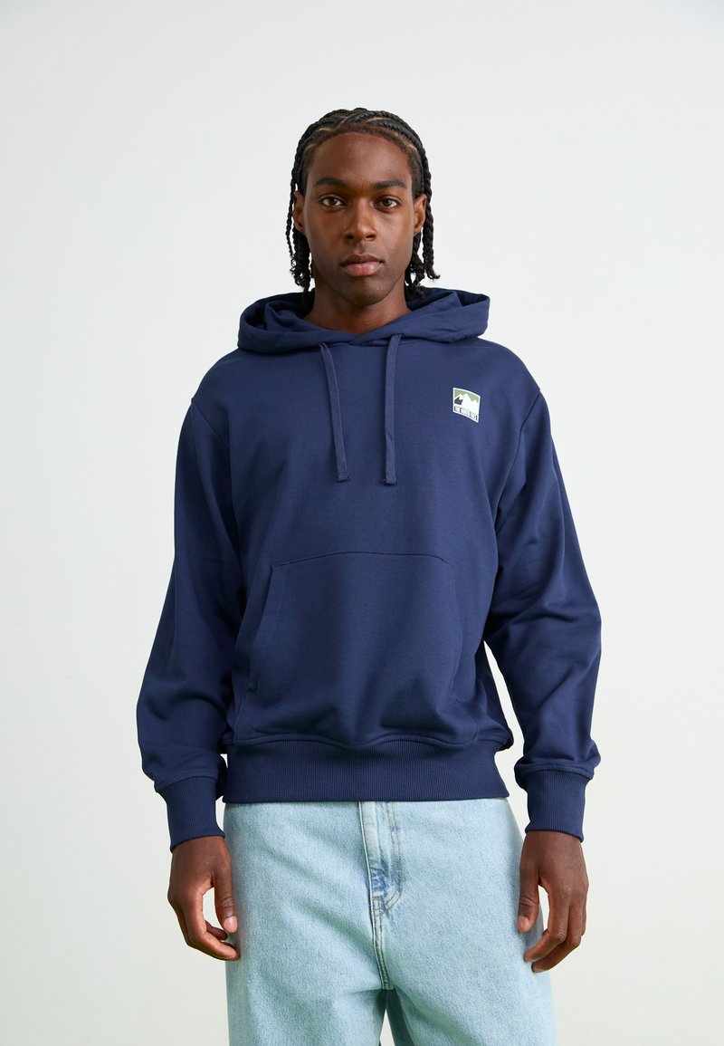 Navy hoodie with a kangaroo pocket and drawstring hood. Features a small mountain logo on the chest. Paired with light blue jeans.