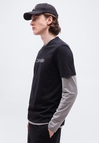 Νεαρός άνδρας με μαύρο καπέλο και μαύρο T-shirt Calvin Klein φορεμένο πάνω από γκρι μακρυμάνικο πουκάμισο, στέκεται σε πλάγια θέση με τα χέρια στις τσέπες.