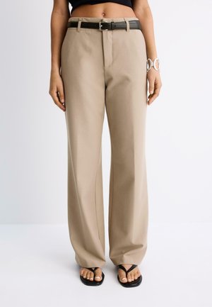 Femme portant un pantalon beige à jambes larges, une ceinture noire, des sandales noires et un bracelet blanc au poignet gauche.