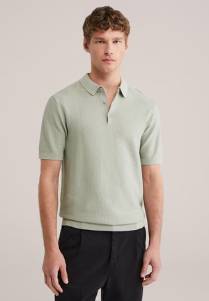 Jonge man met lichtbruin krullend haar, gekleed in een saliegroene, kortärmelig textuurpolo en zwarte broek, staand tegen een neutrale achtergrond.