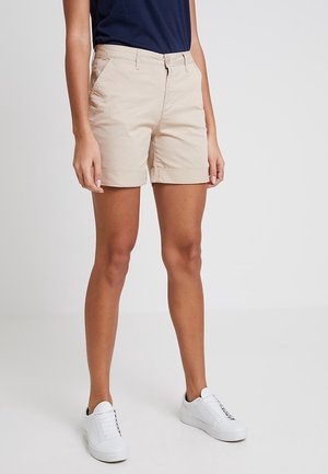 Shorts en coton beige avec un ourlet roulé, dotés de poches latérales et d'une fermeture éclair à l'avant. Associés à des baskets blanches et un haut bleu marine.