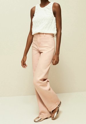 Pantalones anchos color rosa claro de tiro alto, combinados con una blusa sin mangas blanca con textura. La modelo lleva zapatos planos metálicos. Fondo sencillo.