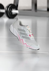Scarpa da corsa grigio chiaro con tomaia in mesh, accenti rosa, suola con motivo a zigzag e tre strisce grigie sul lato. Logo circolare vicino ai lacci.