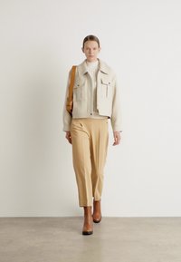 Beige cropped jakke med to brystlommer, cremefarvet sweater, beige skræddersyede bukser og brune ankelstøvler, model går mod en ensfarvet baggrund.