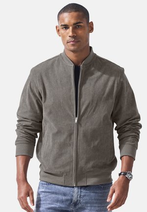 Mann trägt eine graue Cordjacke mit Reißverschluss über einem schwarzen Shirt und blauen Jeans, steht mit entspannten Armen und trägt eine Armbanduhr.