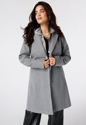 Femme aux longs cheveux foncés portant un manteau gris superposé et un pantalon noir, regardant vers sa gauche sur un fond blanc uni.