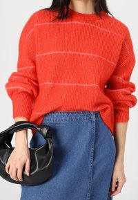 Punainen raidallinen neule, jossa on teksturoitu neulos, pariutettuna korkeavyötäröisen farkkuhameen kanssa; pidetään mustaa, pehmeää tekonahkalaukkua.