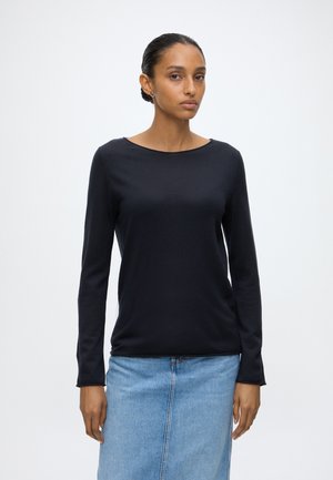 Vrouw met donker haar naar achteren gebonden, gekleed in een zwart top met lange mouwen en een lichtblauwe spijkerrok tegen een effen witte achtergrond.