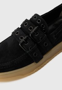 Chaussures en daim noir avec une semelle en caoutchouc beige, des sangles à double boucle avec œillets, et une silhouette épurée avec des détails de couture minimaux.