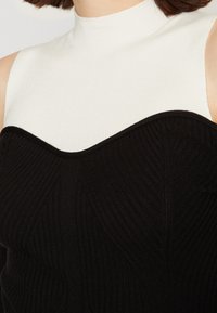 Corpetto nero a coste con scollo strutturato a forma di chevron; sezione bianca a collo alto che contrasta per texture e colore.