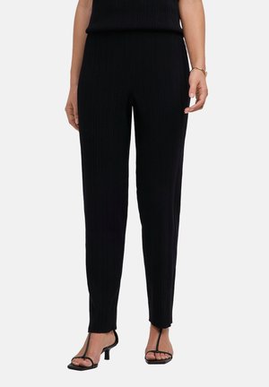 Femme portant un pantalon noir côtelé ajusté avec un haut assorti et des sandales à talons noires à bouts ouverts sur un fond uni.