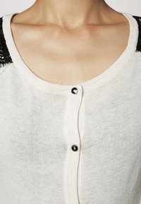 Ljus krämfärgad cardigan gjord av ett mjukt stickat material. Har svarta spetsdetaljer på axlarna, fem knappar framtill och rund halsringning.