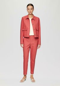 Korallfarbener Blazer mit Knopfleiste und zwei Brusttaschen, kombiniert mit passenden schmal geschnittenen Hosen und weißen Riemchensandalen. Glatte Stofftextur.