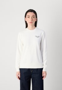 En kvinne står mot en nøytral bakgrunn, iført en hvit genser med marineblå tekst og mørke jeans, og ser direkte på betrakteren.