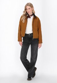 Bruine cropped jack met zwarte kraag, witte shirt, zwarte jeans en zwarte laarzen. Opmerkelijke details zijn onder andere een borstzak en gestructureerde stof.