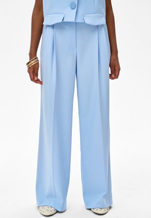 Jasnoniebieskie szerokie spodnie w połączeniu z pasującą marynarką, złote bransolety na lewym nadgarstku oraz białe buty ozdobione ćwiekami na białym tle.