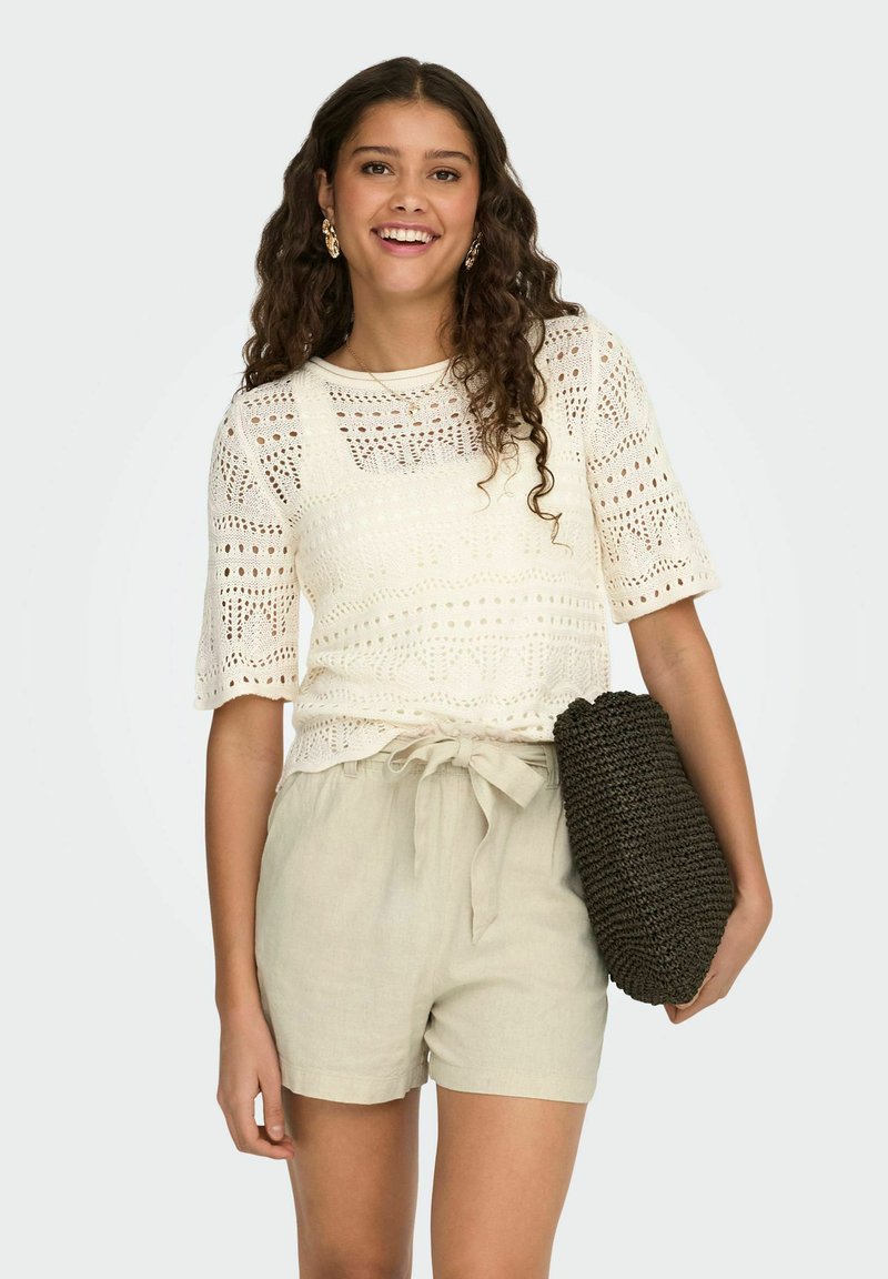 Haut en maille crème à manches courtes, avec un motif ajouré, associé à un short en lin beige clair et un sac texturé foncé.