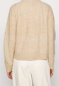 Pull en maille beige avec un ourlet et des poignets côtelés, orné de motifs en côtes texturés courant verticalement le long du dos. Matière douce.