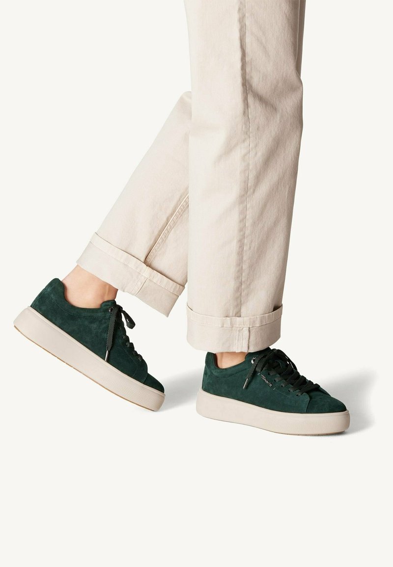Personne portant un pantalon beige et des baskets en daim vert foncé avec des semelles blanches épaisses, debout sur un fond clair uni.