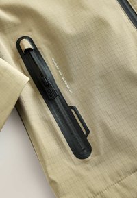 Gros plan d'une veste en tissu beige avec une fermeture éclair noire imperméable et des coordonnées blanches imprimées près de la fermeture éclair.