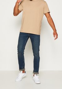 Beige bomull t-shirt, blå slim-fit jeans och vita sneakers. Jeansen har en uppvikt fåll, vilket ger ett avslappnat utseende och en prydlig passform.