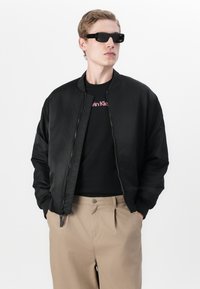 Zwarte bomberjack met rits, gecombineerd met een zwart T-shirt met rode logo-tekst. Beige broek en donkere zonnebril maken de look af.