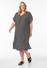 Schwarzes Kleid mit Diamantmuster, kurze Ärmel, V-Ausschnitt, knielang, leichtes Material. Gestylt mit schwarzen Sandalen mit Ausschnitten.