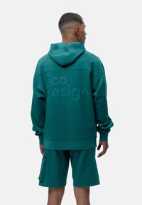 Tyrkysová ekologická mikina s mäkkou textúrou, so kapucňou a výrazným textom "eko dizajn" na chrbte. V komplete s vyhovujúcimi šortkami.
