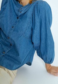 Džínová košile ve středně modré barvě s knoflíkovou légou vpředu, nafouklými rukávy, náprsným detailem a nařasenými manžetami. Měkká textura s pohodlným střihem.