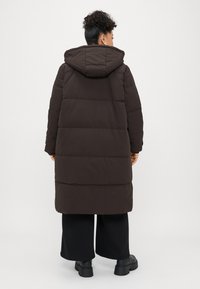 Brown puffy coat with a hood, featuring quilted sections, a mid-length design, and a smooth texture. Worn over black wide-leg pants.