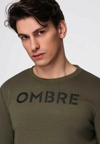 Bluza w kolorze oliwkowej zieleni z okrągłym dekoltem, z dużym czarnym napisem "OMBRE" na piersi. Gładka faktura i dopasowany krój.