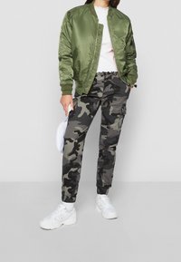 Chaqueta bomber de satén verde olivo sobre una blusa blanca, combinada con pantalones cargo de camuflaje gris y zapatillas blancas. Las características clave incluyen puños elásticos y bolsillos.