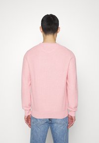 Pull rose pâle avec un tricot texturé, col ras du cou, poignets et ourlet côtelés. Porté avec un jean en denim bleu, présenté de dos.
