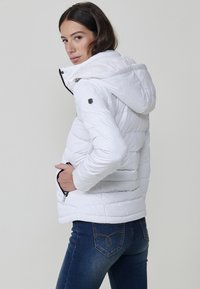 Chaqueta acolchada blanca con capucha, diseño acolchado, bolsillos con cremallera y detalles en negro. Llevada sobre jeans azules, mostrando una silueta ajustada.