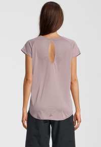 Kobieta w różowej bluzce z krótkim rękawem z wycięciem z tyłu w kształcie klucza i czarnych szortach, stojąca na jednolitym jasnym tle.