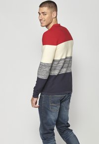 Maglione a blocchi di colore con strisce rosse, crema, grigie e blu navy, texture a coste, maniche lunghe e scollo rotondo. Indossato con jeans blu.