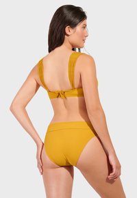 Žuti bikini s teksturiranim debelim trakama i dizajnom vezivanja na leđima. Donji dio ima visoki struk, u boji i materijalu koji odgovara gornjem dijelu.