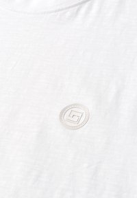 Gros plan sur un tissu blanc avec une petite pièce ronde présentant un logo géométrique cousu près du centre.