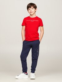 Sarkans kokvilnas T-krekls ar baltu "TOMMY HILFIGER EST. 1985" logotipu, apvienots ar tumši zilām sporta biksēm ar tekstu sānos un baltām kedām.