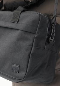Geantă neagră texturată crossbody cu buzunar frontal, mâner superior, închidere cu fermoar și curea de umăr reglabilă, așezată pe un fundal din material textil închis la culoare.